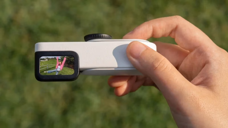 Image 3: A hand holds a small, rectangular camera with a display screen showing a person in a pink shirt doing a handstand on grass, with a blurred green background.